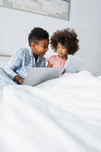 Irmãos afro-americanos usando laptop e tablet digital na cama — Fotografia de Stock