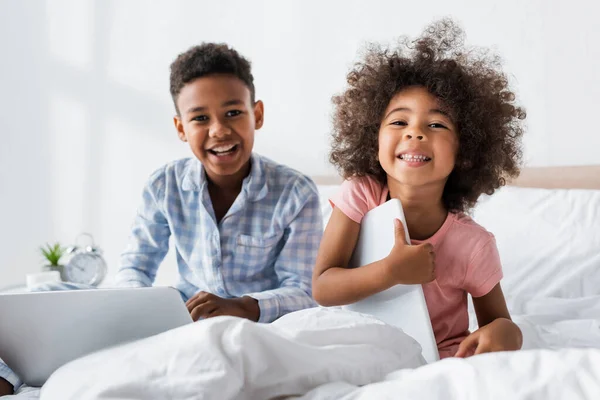 Niños afroamericanos emocionados mirando la cámara mientras están sentados con gadgets en la cama - foto de stock
