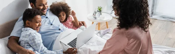 Niños afroamericanos felices mirando el ordenador portátil cerca de papá y la madre borrosa en el dormitorio, pancarta - foto de stock