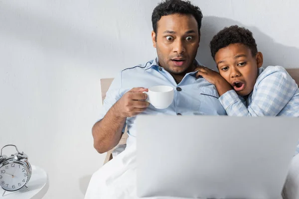 Shocked african american father and son watching movie on laptop in bed — Stock Photo
