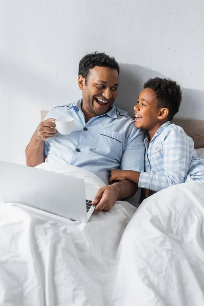 Lachend schauen sich Vater und Sohn in der Nähe des Laptops im Bett an — Stockfoto