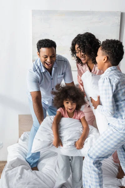 Familia afroamericana emocionada en pijama divirtiéndose con almohadas en el dormitorio - foto de stock