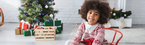 Glückliches afrikanisch-amerikanisches Mädchen blickt in die Kamera, während es auf einem Schlitten neben verschwommenen Geschenken unter dem Weihnachtsbaum sitzt, Banner — Stockfoto