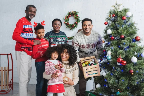 Uomo in possesso di scatola di legno con palline decorative vicino all'albero di Natale e famiglia afro-americana — Foto stock