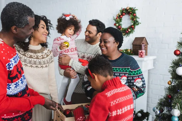 Niño sosteniendo caja de madera con bolas de Navidad cerca de alegre familia afroamericana en la sala de estar - foto de stock