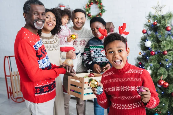 Aufgeregten afrikanisch-amerikanischen Jungen in Rentierhörnern Stirnband hält Weihnachtskugeln in der Nähe glücklicher Familie — Stockfoto