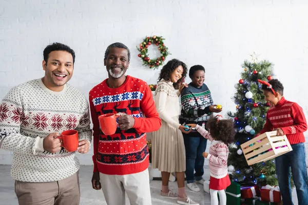 Cheerful men with cups near blurred african american family decorating christmas tree — Stock Photo