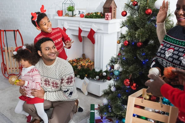 Emocionado afroamericano chico saludando de la mano a borrosa abuelos decorando árbol de Navidad cerca de hombre con hija - foto de stock