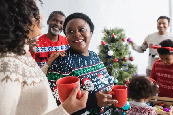 Afro-américaine femmes avec des tasses souriant à l'autre près floue famille décorer arbre de Noël — Photo de stock
