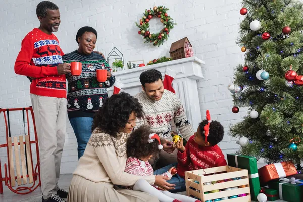 Padres afroamericanos con niños eligiendo adornos decorativos cerca del árbol de Navidad y abuelos con copas — Stock Photo