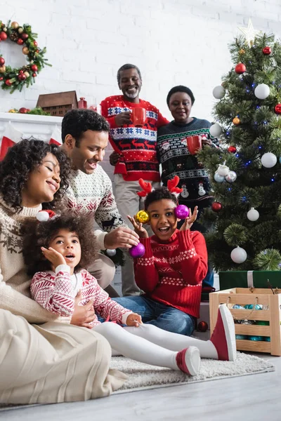 Excited boy in reindeer horns headband holding decorative baubles near christmas tree and smiling african american family — Stock Photo