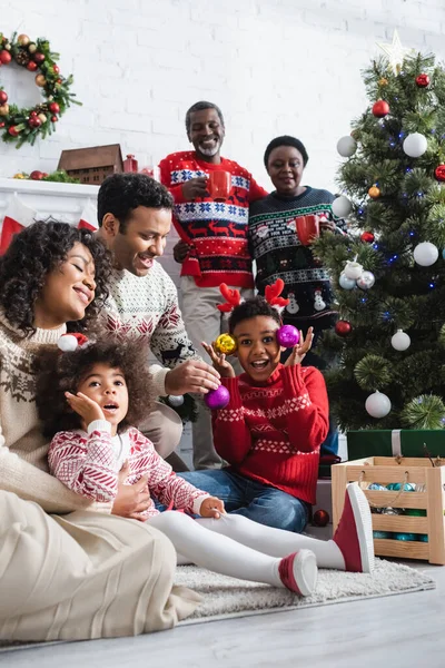 Menino animado em chifres de rena headband segurando bugigangas decorativas perto de árvore de natal e família afro-americana sorridente — Fotografia de Stock