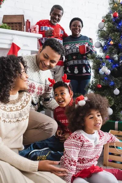 Alegre afroamericano familia cerca decorado árbol de Navidad y borrosa abuelos con tazas — Stock Photo
