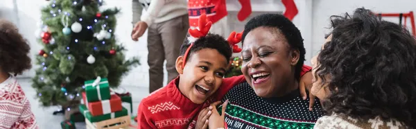 Feliz chico y mujer abrazando riendo africano americano abuelita cerca borrosa familia y árbol de navidad, bandera - foto de stock