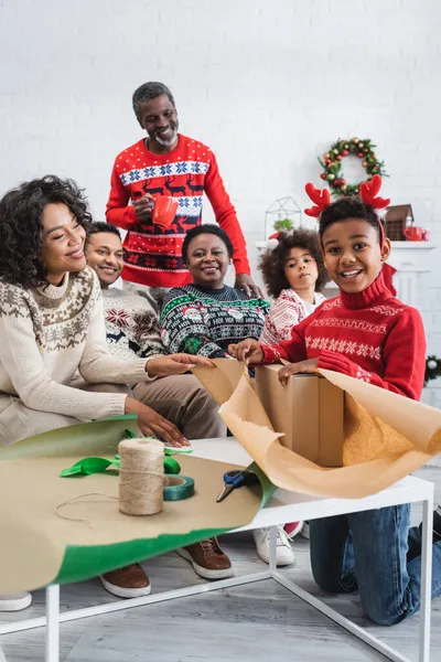 Cheerful boy in reindeer horns headband packing christmas present near happy african american family — Stock Photo