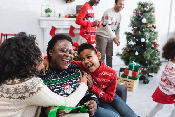 Feliz chico y mujer abrazando riendo africano americano abuelita cerca borrosa hombres y árbol de Navidad en sala de estar - foto de stock