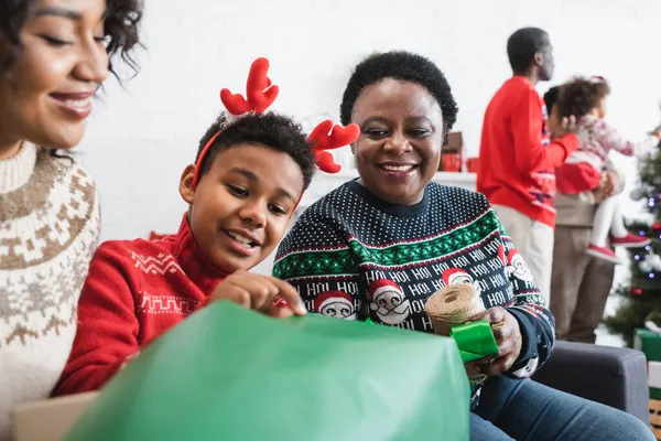 Africano americano chico en renos cuernos diadema embalaje navidad presente con complacido madre y abuelita - foto de stock