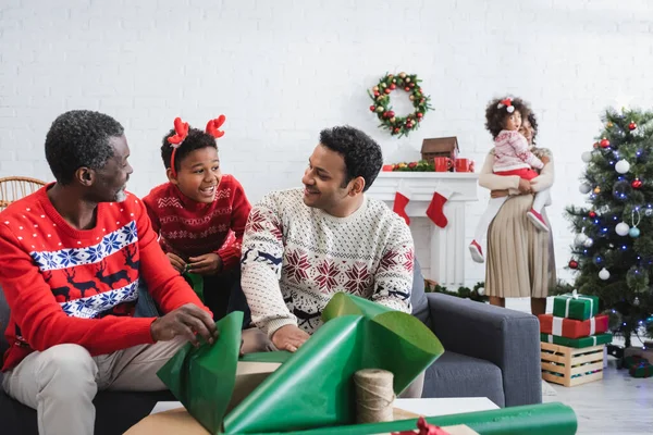Африканский американский мальчик в оленьих рогах ободок улыбается рядом с мужчинами, упаковывая подарки и размытая мать с ребенком возле рождественской елки — стоковое фото