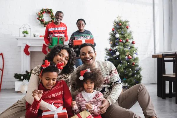 Familia afroamericana feliz con cajas de regalo mirando a la cámara cerca de los abuelos borrosos y el árbol de Navidad - foto de stock