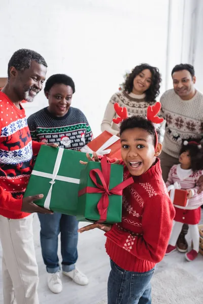 Amazed african american boy in headband with reindeer horns showing christmas present near blurred family — Stock Photo