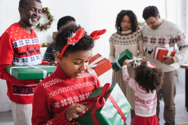 Amazed african american boy in reindeer horns headband holding xmas present near blurred family with gift boxes — Stock Photo