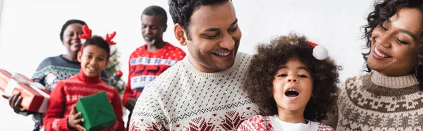 Усміхнені батьки біля здивованої дочки і розмита афроамериканська сім'я з різдвяними подарунками, банер — стокове фото