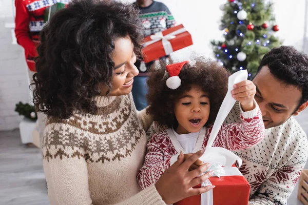 Menina americana africana espantada abrindo presentes de Natal perto de pais felizes e árvore de Natal borrada — Fotografia de Stock
