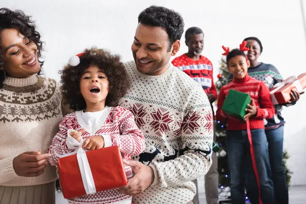 Happy parents near amazed daughter with gift box and african american family on blurred background — Stock Photo