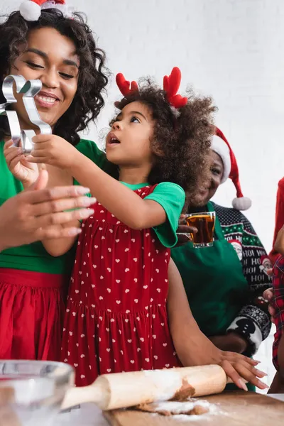 Africano americano chica en renos cuernos diadema celebración cookie cutter cerca feliz mamá y sonriente abuelita - foto de stock