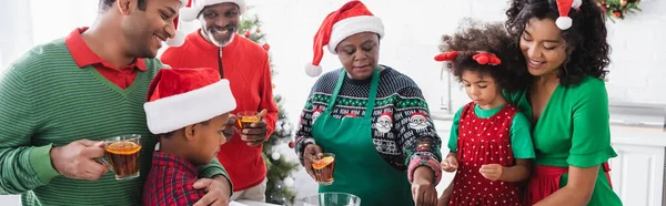 Famiglia afroamericana in cappelli di Babbo Natale sorridente vicino nonna cucina in cucina, banner — Foto stock