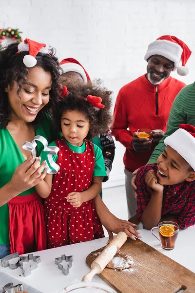 Femme afro-américaine montrant coupe-biscuits à la fille près de la pâte et la famille heureuse avec du thé à la cannelle orange — Photo de stock