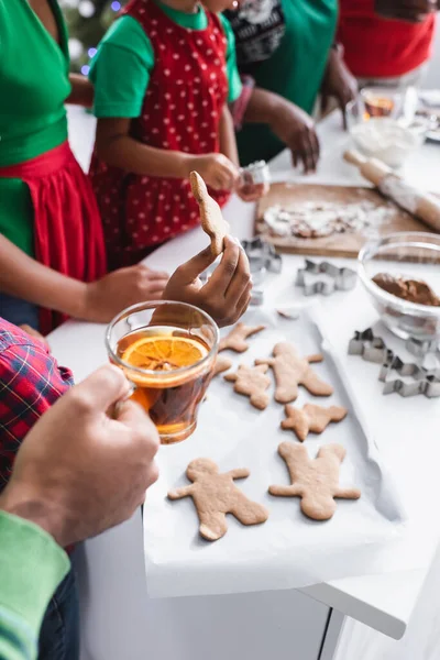 Vista recortada de niño afroamericano con galleta de Navidad horneada cerca del padre con té de canela naranja y familia borrosa - foto de stock