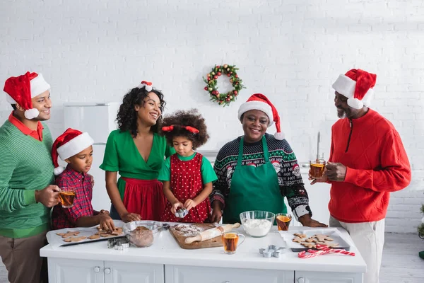 Счастливые африканские американские мужчины с чашками апельсинового чая с корицей рядом с женщинами и детьми, готовящими рождественское печенье — стоковое фото