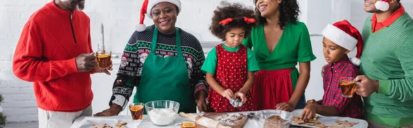 Счастливые африканские американские женщины и дети, готовящие рождественские печенья рядом с мужчинами, пьющие апельсиновый чай с корицей, баннер — стоковое фото