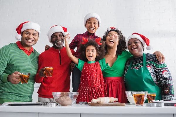 Afroamericana chica cantando cerca feliz familia en santa sombreros y los ingredientes en mesa - foto de stock