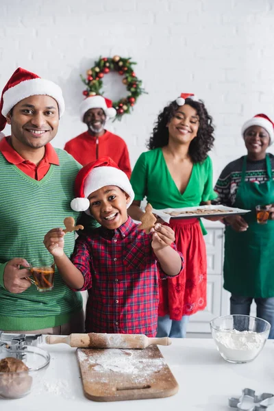 Alegre menino afro-americano mostrando cozido biscoito de Natal perto da família em chapéus de santa na cozinha — Fotografia de Stock