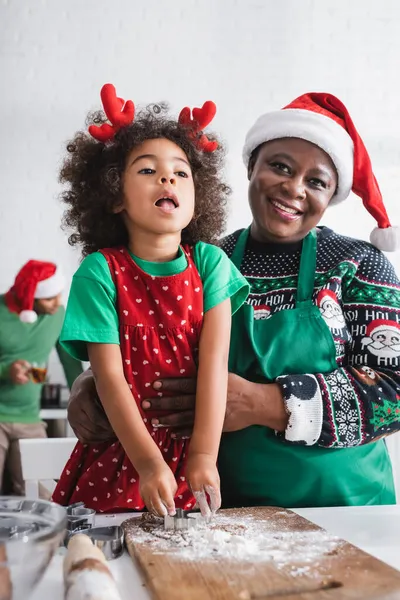 Glücklich afrikanisch-amerikanische Frau mit Weihnachtsmannmütze umarmt Enkelin beim gemeinsamen Kochen in der Küche — Stockfoto