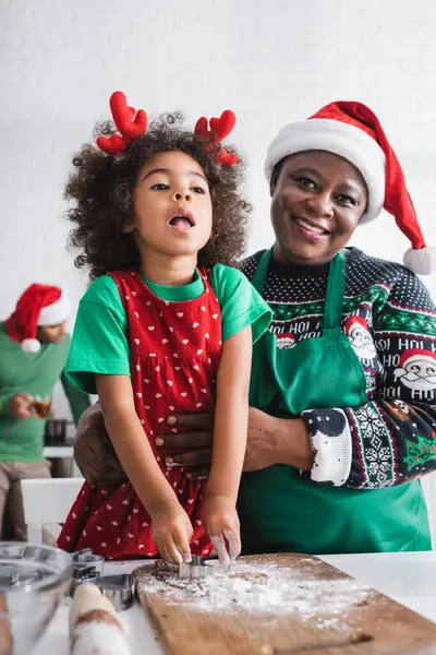 Feliz africana americana mujer en santa hat abrazando nieta mientras cocinar en cocina juntos - foto de stock