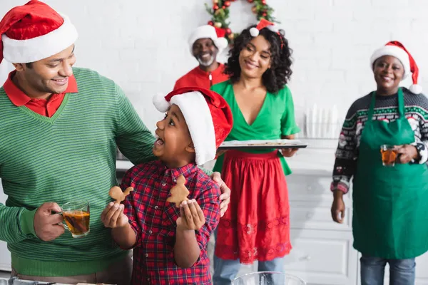 Stupito ragazzo africano americano in cappello di Babbo Natale mostrando biscotti di Natale a papà con tazza di tè alla cannella arancione — Foto stock