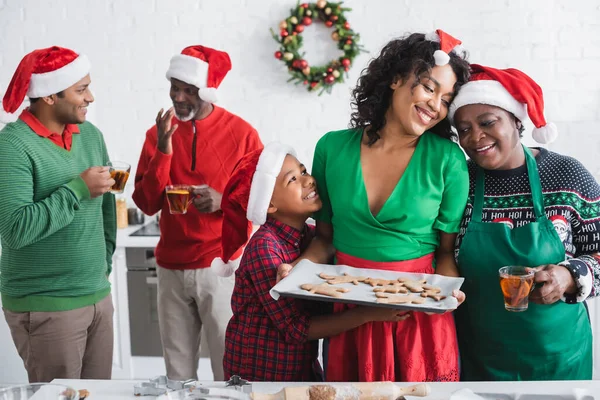 Афроамериканська жінка тримає лист для випічки з різдвяним печивом біля щасливої сім'ї на кухні — стокове фото
