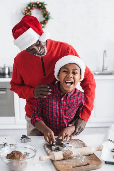 Aufgeregten afrikanisch-amerikanischen Jungen in Weihnachtsmütze Teig schneiden mit Ausstecher in der Nähe glücklichen Opa in der Küche — Stockfoto