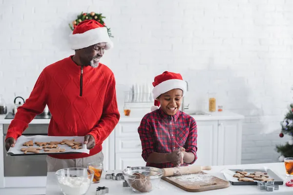 Hombre afroamericano feliz sosteniendo bandeja para hornear con galletas de Navidad cerca de nieto - foto de stock