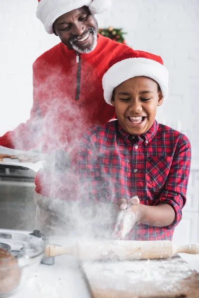 Lächelnder afrikanisch-amerikanischer Mann schaut Enkel mit Weihnachtsmütze in der Nähe von Mehl in der Küche an — Stockfoto