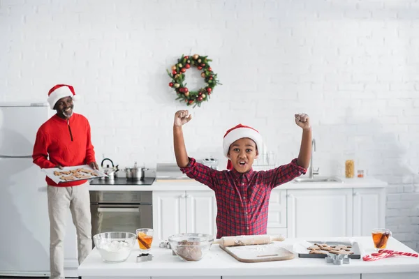 Збуджений афро-американський хлопчик у капелюсі Санти, що показує жест успіху біля розмитого дідуся з сирим різдвяним печивом на кухні — стокове фото