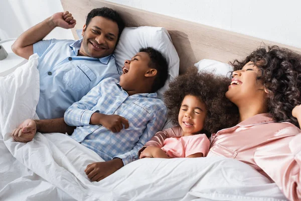 Menino americano africano bocejando de manhã enquanto deitado na cama perto de pais felizes e irmã — Fotografia de Stock