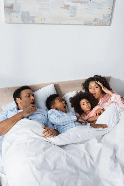 Overslept and shocked african american couple lying with kids in bed — Stock Photo