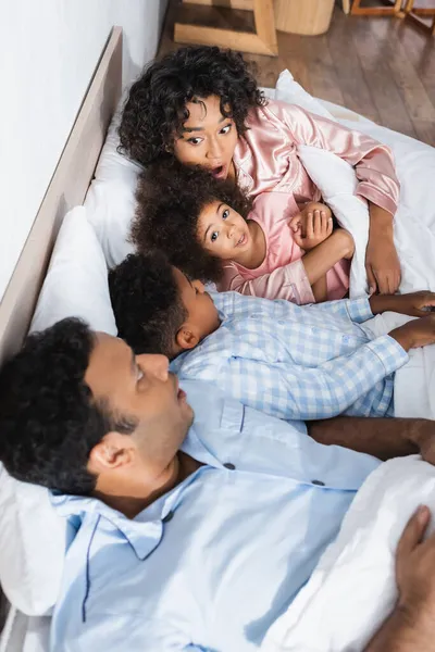 Overslept african american couple looking at each other while lying with kids in bed — Stock Photo