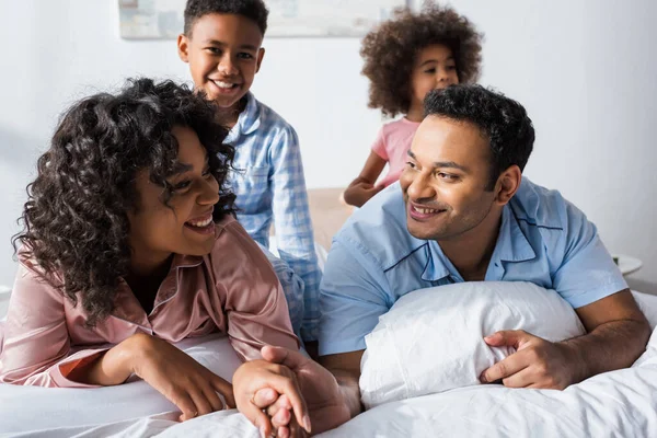 Felice uomo africano americano che tiene la mano della moglie vicino a bambini sfocati in camera da letto — Foto stock