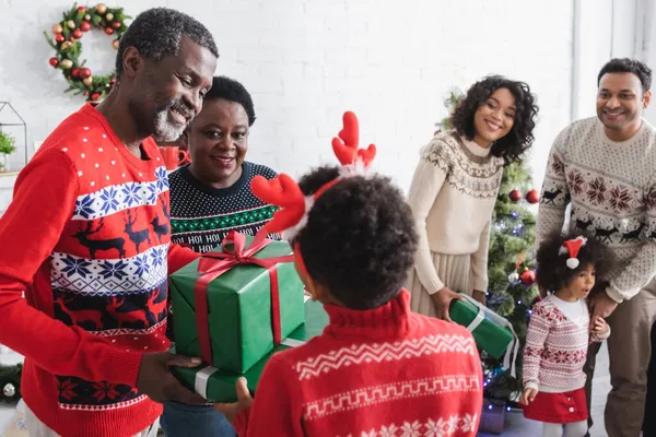 Vista posterior del chico afroamericano en diadema de cuernos de reno presentando regalos de Navidad a abuelos felices - foto de stock