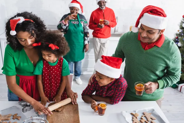 African american woman rolling out dough near family and blurred grandparents drinking orange cinnamon tea — Stock Photo